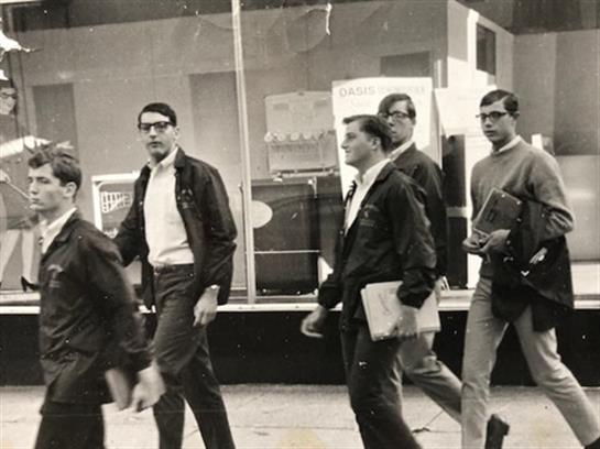 Young men in casual clothes chat as they stroll past shop windows in a vibrant city.
