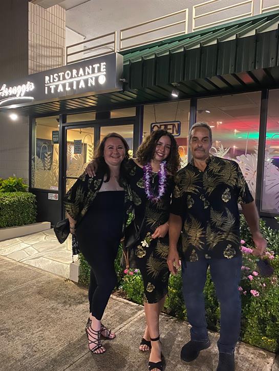 Three friends dressed in floral outfits pose happily outside a restaurant with bright lights.