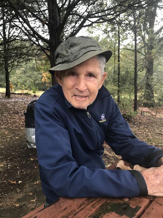 Elderly man wearing a hat relaxes on a bench in a wooded area surrounded by trees.