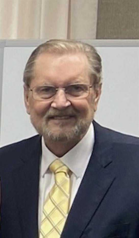 A senior man in formal attire stands and smiles, engaging with others at a gathering.