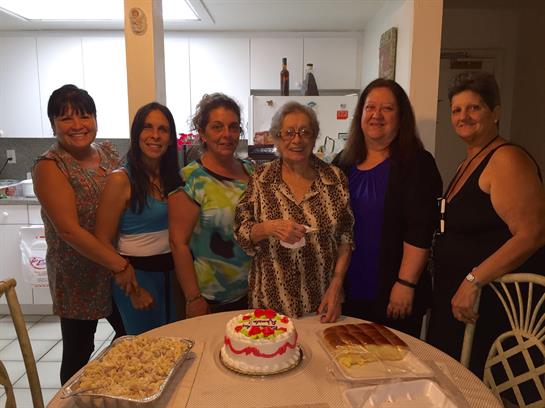 A group of six women celebrate together in a warm kitchen while enjoying a cake and treats.
