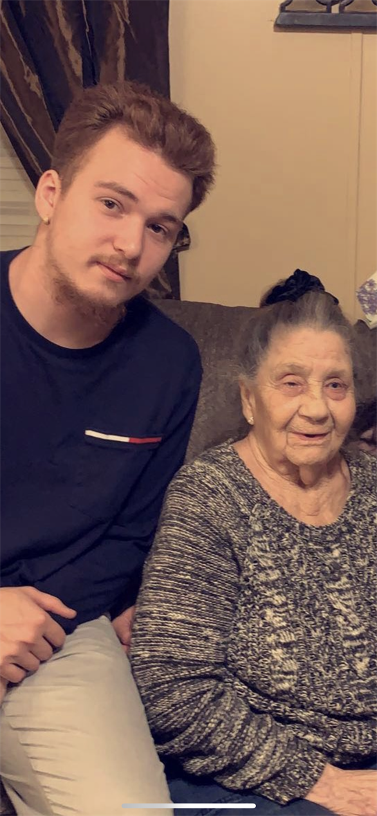 A young man sits next to his grandmother, both smiling warmly, enjoying a family moment indoors.
