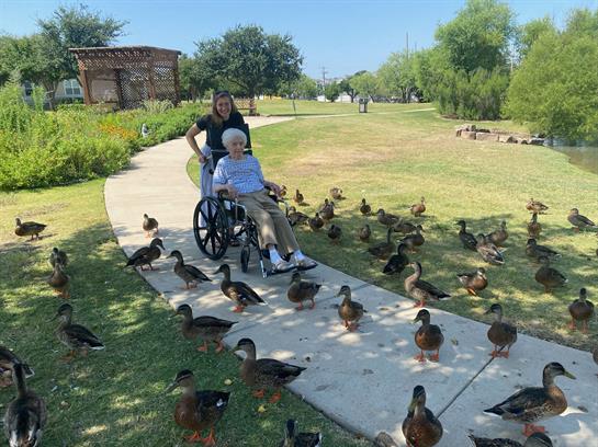 A woman in a wheelchair smiles while a caretaker pushes her along a park path filled with ducks.
