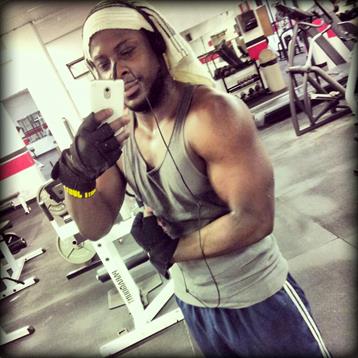 A man flexes his arm while taking a selfie in a gym, surrounded by weights and fitness machines.