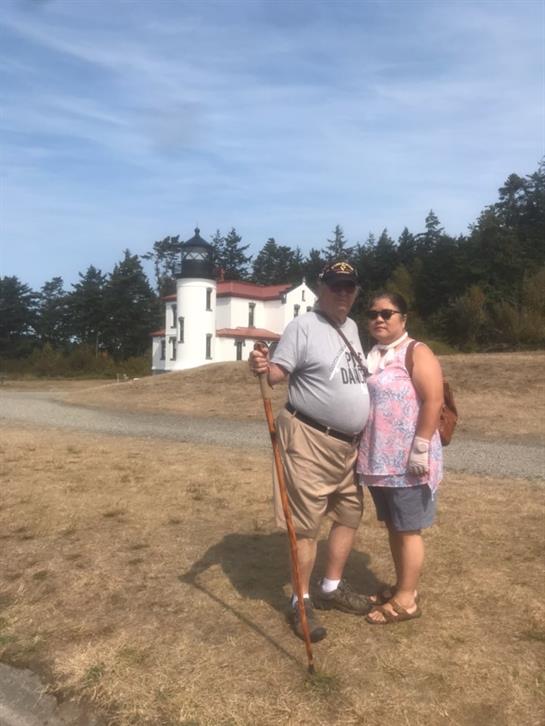 Two people stand together smiling in front of a lighthouse on a clear, sunny day.