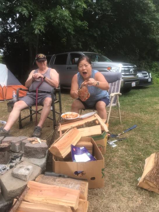 Two friends are seated by a campfire, enjoying barbecue food and sharing laughter outdoors.