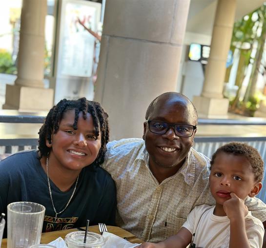 A family gathers around a table, sharing a joyful meal outdoors under warm sunlight.
