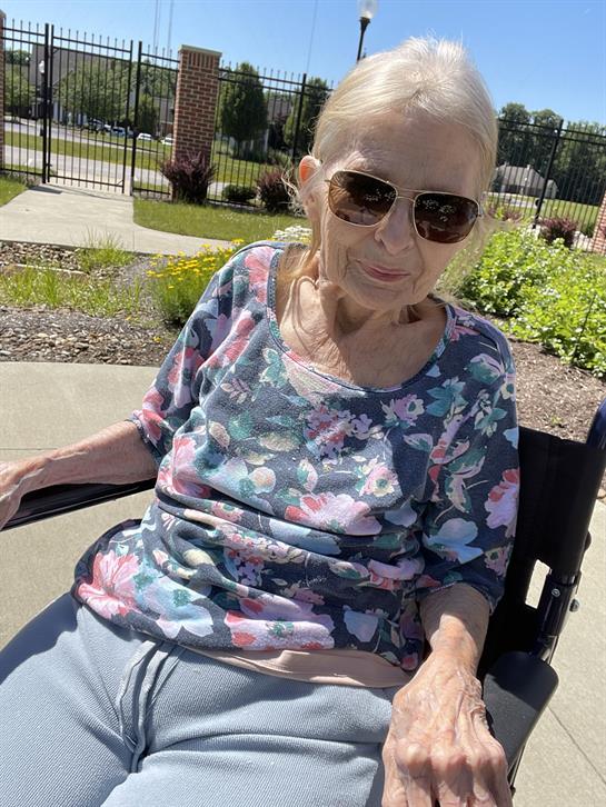 Elderly woman with sunglasses relaxes in her wheelchair, surrounded by greenery and sunshine.
