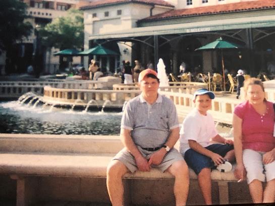 Family relaxes near a fountain, enjoying a sunny day in a lively town square with shops.