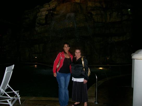 Two friends enjoy a night out, posing together with a pool and rocky backdrop under the stars.