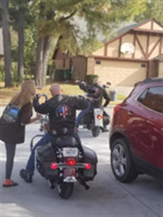 Two motorcyclists converse with a woman near parked cars on a clear day in a residential area.