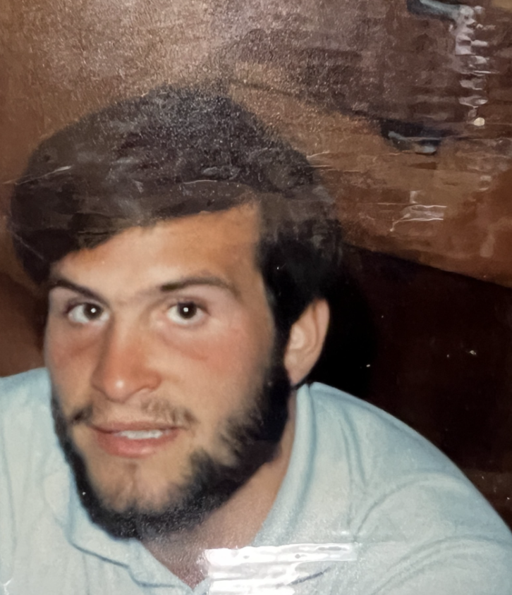 A man with a beard is smiling while sitting close to the camera in a cozy, wooden interior.