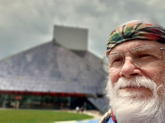 An elderly man smiles happily in a stadium, donning a colorful headband on a sunny day.