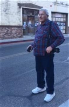 A man with a camera poses in front of a classic structure in a city street under clear skies.