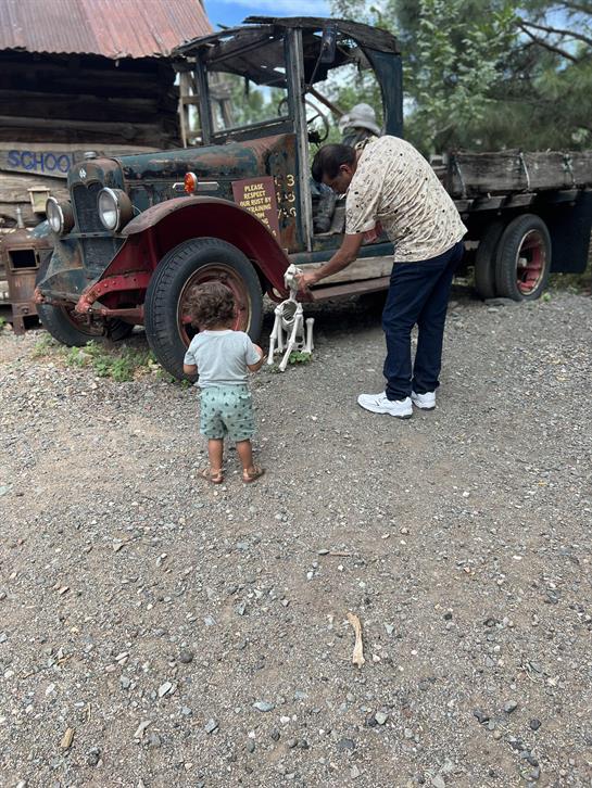 A young child watches as an adult engages with a small dog beside an old truck.