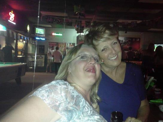 Two friends are smiling and posing together in a bar, enjoying their evening out with drinks.