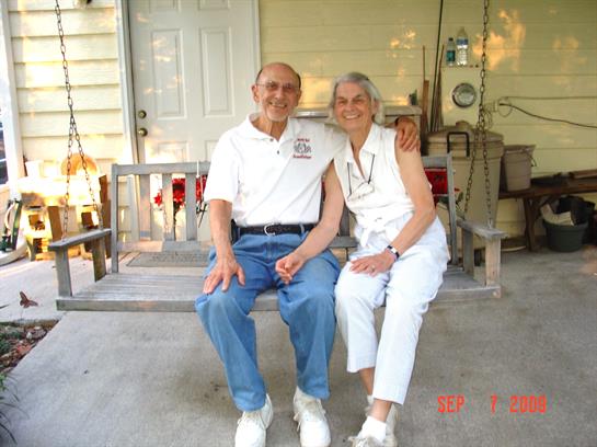 An elderly couple smiles together on a porch swing, enjoying a warm afternoon.
