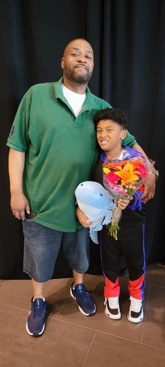 A boy joyfully holds a bouquet of flowers and a stuffed dolphin, standing next to an adult.