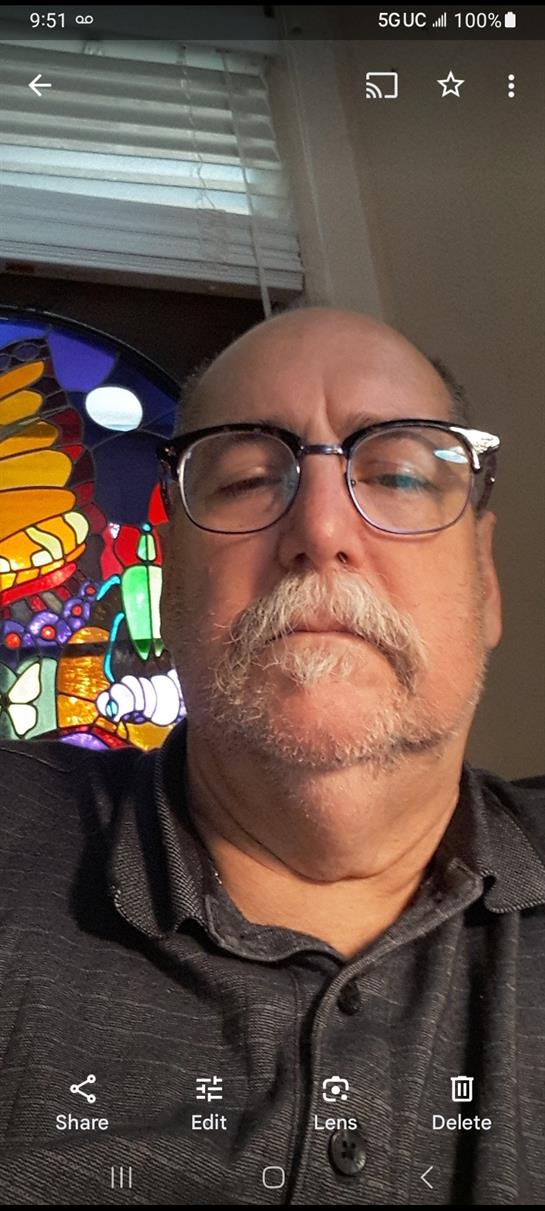 Man with glasses poses for a self-portrait near a vibrant stained glass window at home.