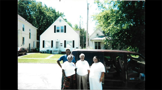 Four family members stand together smiling outside a house, enjoying a sunny day.