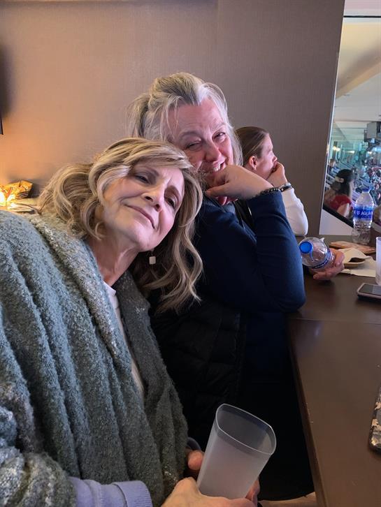 Two women share a moment of laughter at a bar wrapped in cozy blankets while others sip drinks.