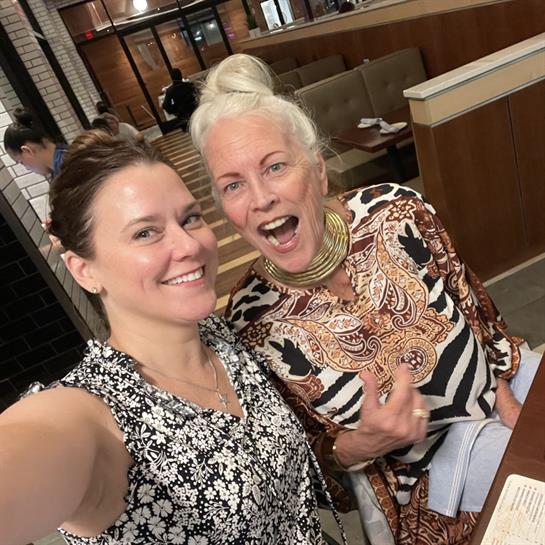 Two women smile and pose for a cheerful selfie while enjoying their time in a busy cafe.