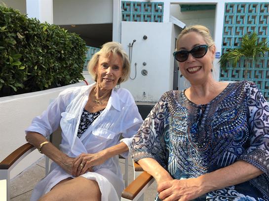 Two women relax by the poolside, dressed casually, enjoying the sunny weather in a tropical setting.