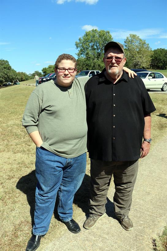 Two men smile while posing for a picture on a clear day, surrounded by parked cars in nature.