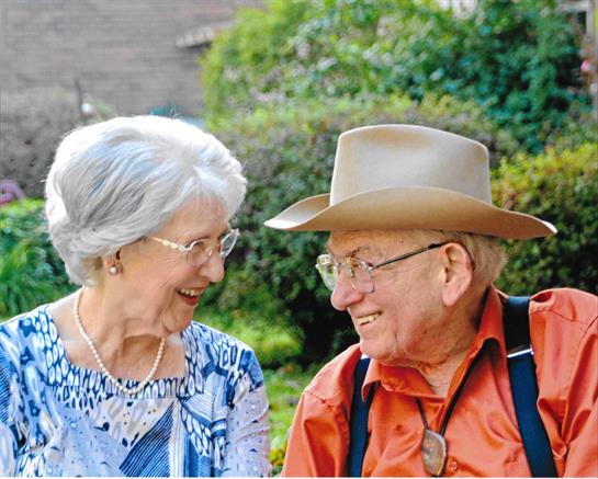 Two seniors share a warm smile and laughter in a vibrant garden on a clear day.
