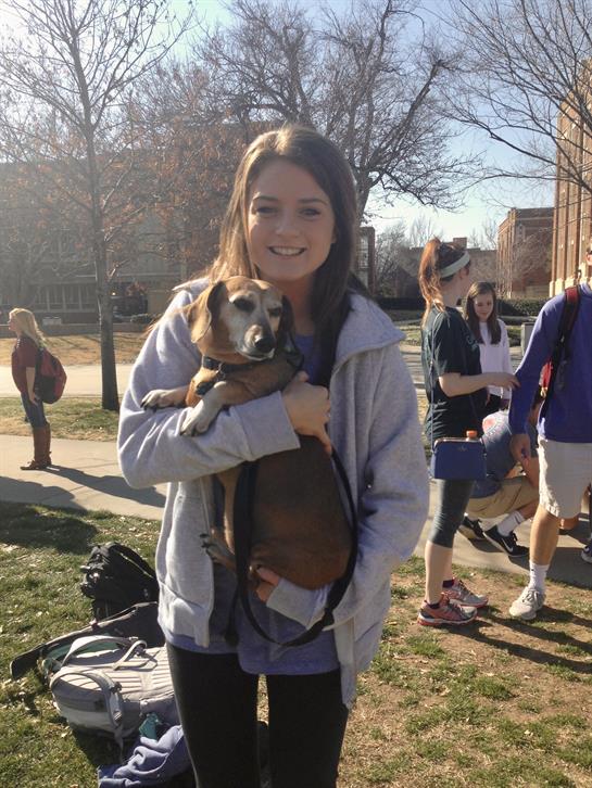 A young woman joyfully holds a small brown dog while surrounded by fellow students on campus.