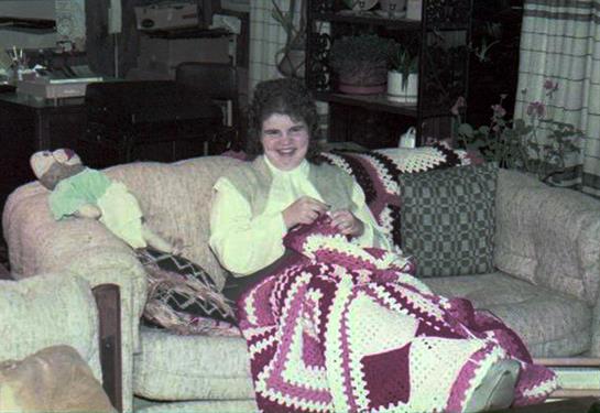 A woman knits a colorful blanket on a sofa in a cozy, craft-filled living room.
