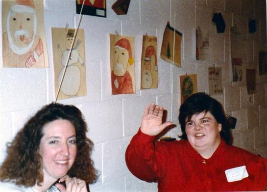 Two women smile and wave in front of colorful children's artwork displayed on a wall.