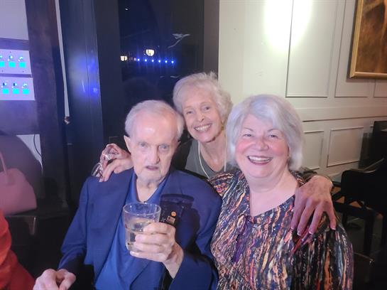 Three elderly friends share joyful moments and drinks at a cozy evening gathering.