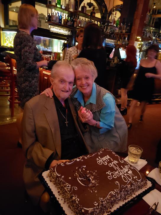 An elderly man enjoys his birthday party with a woman at a restaurant, surrounded by loved ones.