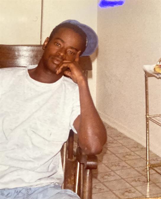 A young man rests his chin on his hand, reflecting quietly in a modest room with tiled floor.