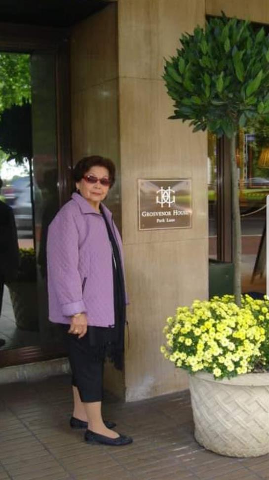 A woman wearing sunglasses and a purple jacket poses by hotel doors surrounded by flowers.