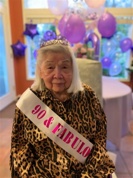Elderly woman wears a tiara and sash at a joyful birthday celebration surrounded by decorations.