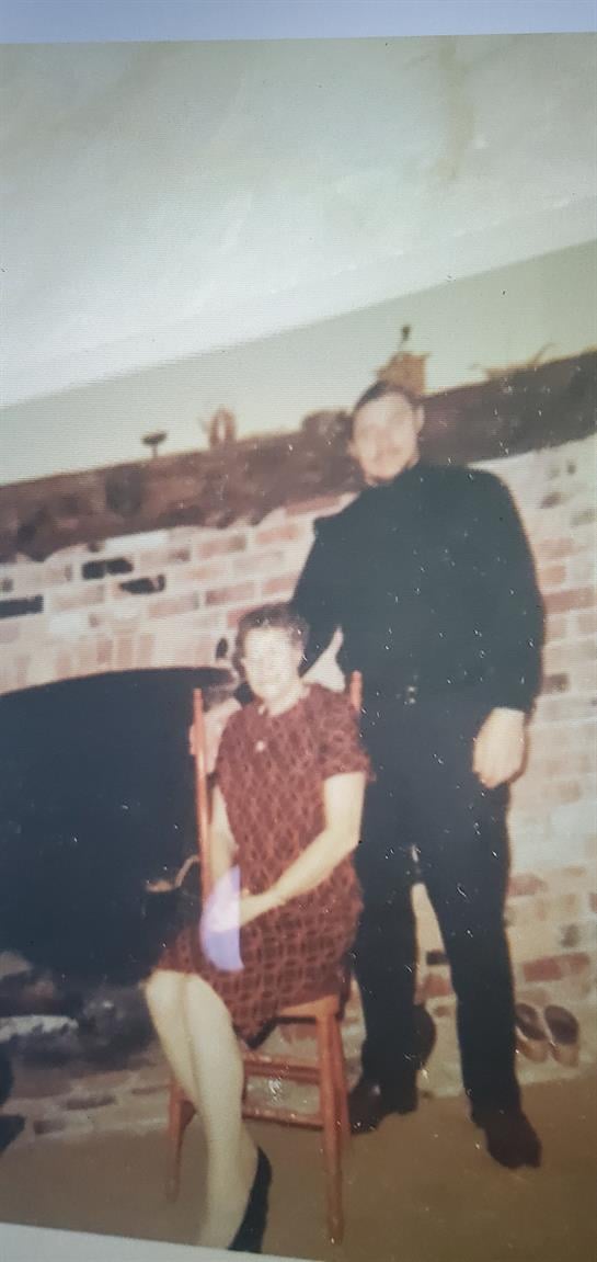 A man stands beside a woman seated in front of a brick fireplace, enjoying a warm moment.