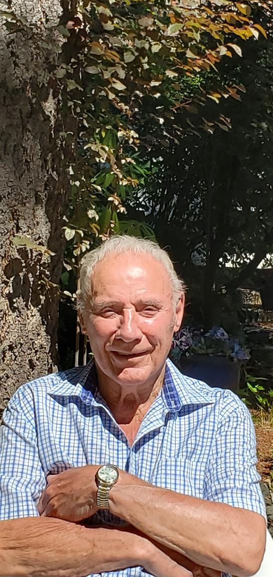 A cheerful elderly man stands outside surrounded by lush green foliage on a bright day.