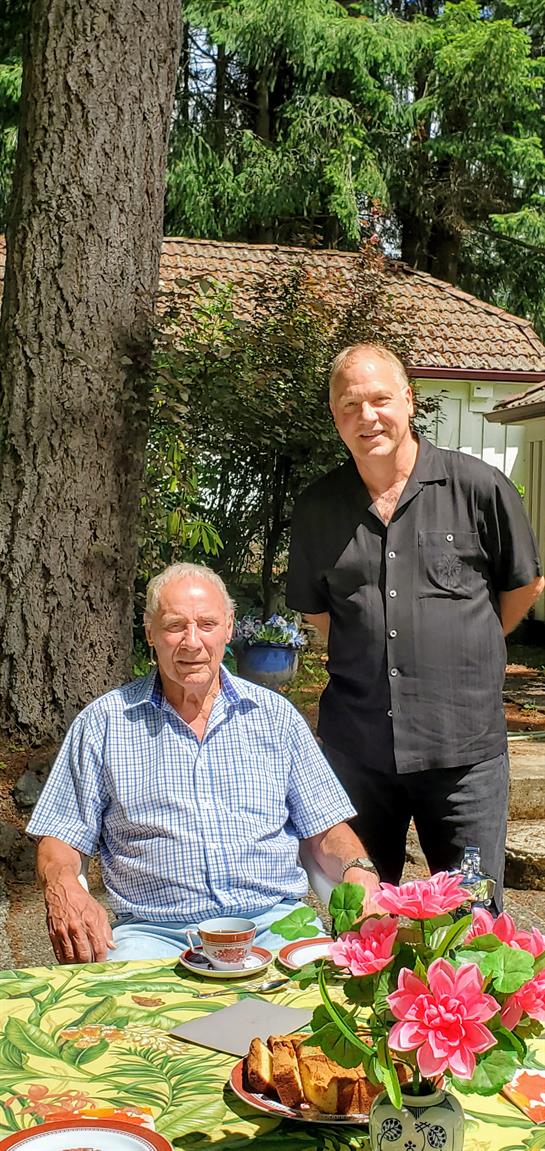 Two men are enjoying a relaxed moment outdoors in a garden on a clear, sunny day.