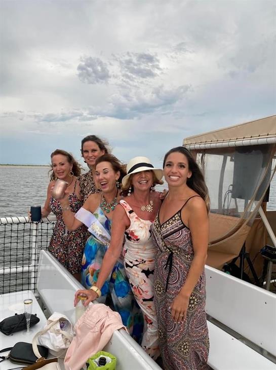 Five women celebrate together on a boat under an overcast sky, smiling and enjoying.