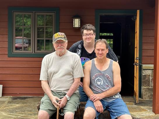Three men sit outside a rustic cabin, enjoying a sunny summer day and sharing moments together.