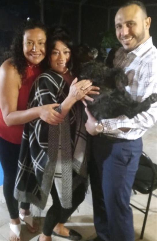 Two women and a man joyfully embrace one another while holding a dog in a warm indoor atmosphere.