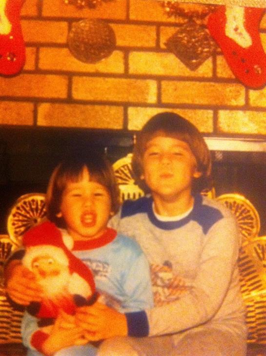 Two young boys smile while holding a Santa doll, sitting by a festive fireplace during Christmas.