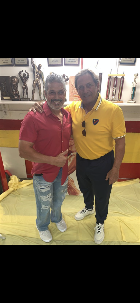 Two men stand close, smiling and posing with a thumbs-up in an artful space.