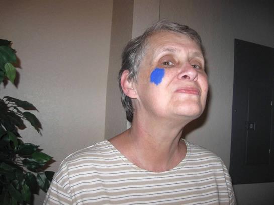 A woman with gray hair shows a playful smile, sporting blue paint on her cheek in a cozy room.