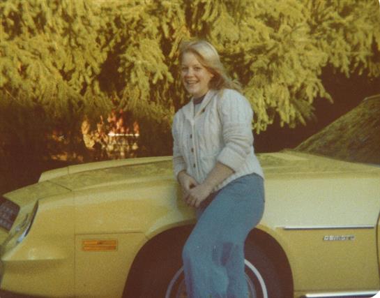 A woman smiles while leaning against a yellow vintage car surrounded by green foliage.