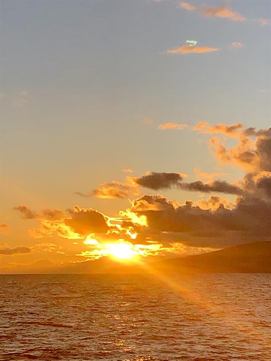 Sunlight breaks through clouds as it sets over the tranquil ocean, painting the sky warm shades.