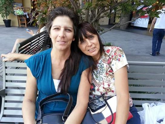 Two women sit closely on a bench, smiling and enjoying each other's company on a sunny day.
