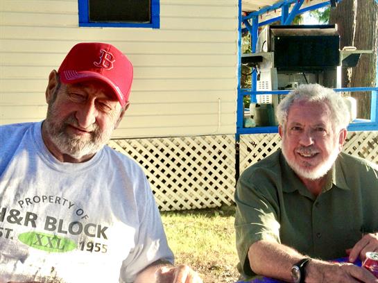 Two men smile while seated at a table, enjoying food in a sunny outdoor gathering.